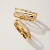 The Nola Pavé Triple Band Ring in Gold features three overlapping, pavé diamond-encrusted bands, elegantly displayed on a light beige surface with soft shadows.