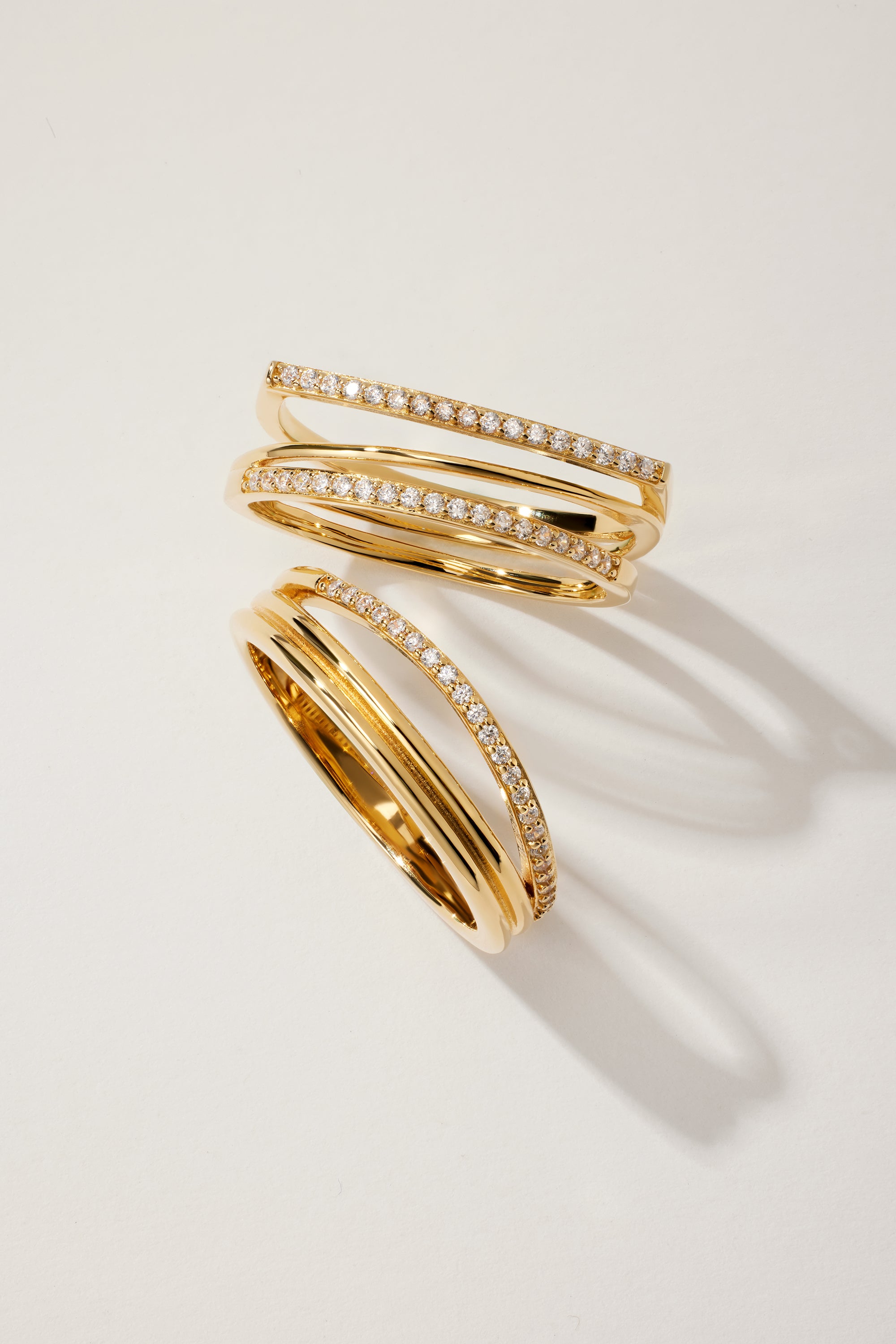 The Nola Pavé Triple Band Ring in Gold features three overlapping, pavé diamond-encrusted bands, elegantly displayed on a light beige surface with soft shadows.