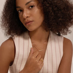 A woman with curly hair wears a sleeveless, light-striped top and minimalist jewellery, including the Regina Horizontal Bar Pendant Necklace in Gold, along with cubic zirconia rings, posing against a neutral background.