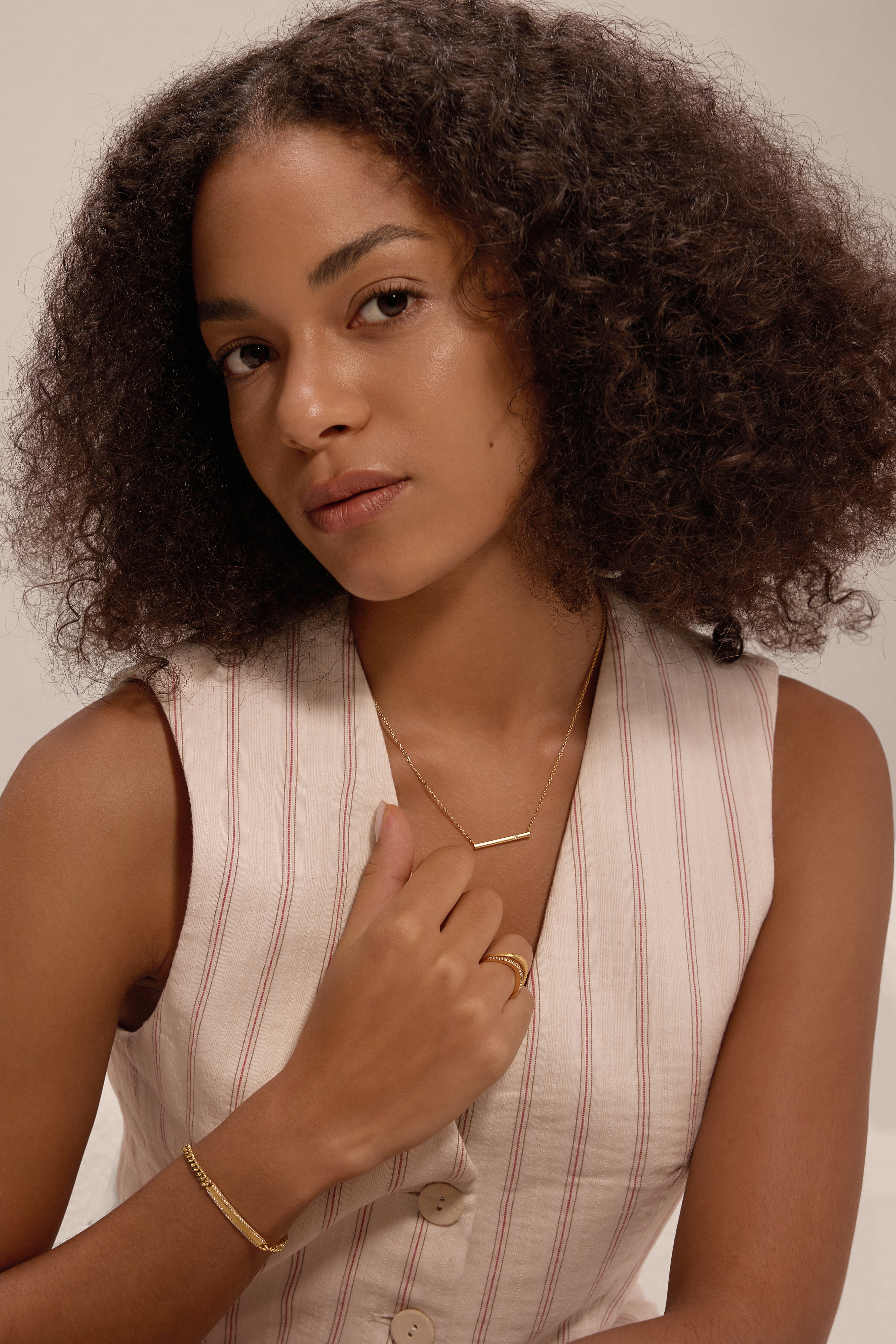 A woman with curly hair wears a sleeveless, light-striped top and minimalist jewellery, including the Regina Horizontal Bar Pendant Necklace in Gold, along with cubic zirconia rings, posing against a neutral background.