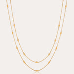 The Simona Double Chain Stacking Necklace in Gold features two delicate chains with evenly spaced oval and round bead accents, making it a minimalist piece thats perfect for layering or wearing solo against a light background.