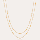 The Simona Double Chain Stacking Necklace in Gold features two delicate chains with evenly spaced oval and round bead accents, making it a minimalist piece thats perfect for layering or wearing solo against a light background.