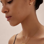 A woman with her dark hair tied back wears the Judith Hexagon Angular Huggies in Gold and a delicate gold bar necklace, dressed in a black strap top, posing against a neutral background.