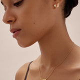 A woman with her dark hair tied back wears the Judith Hexagon Angular Huggies in Gold and a delicate gold bar necklace, dressed in a black strap top, posing against a neutral background.