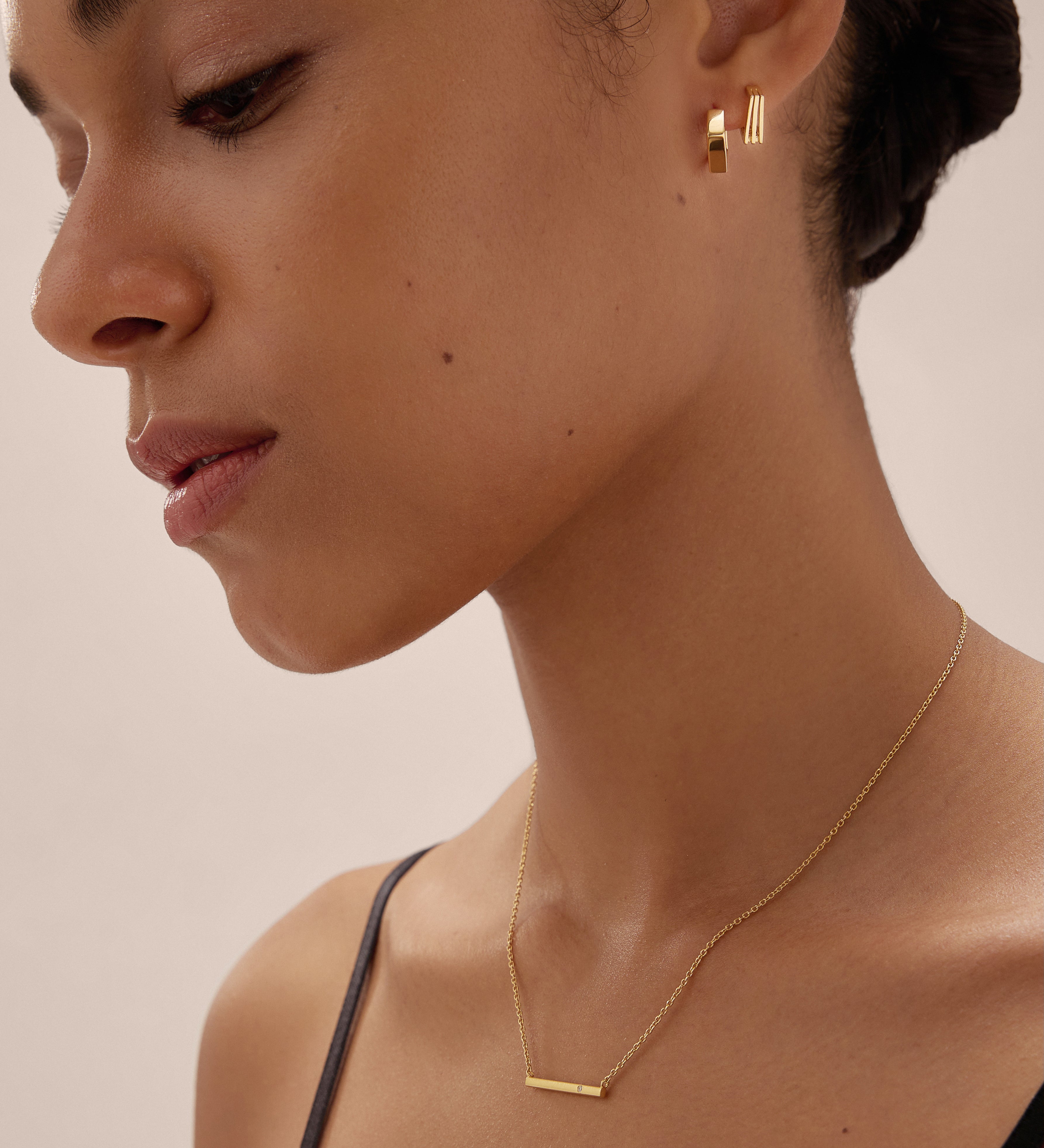 A woman with her dark hair tied back wears the Judith Hexagon Angular Huggies in Gold and a delicate gold bar necklace, dressed in a black strap top, posing against a neutral background.