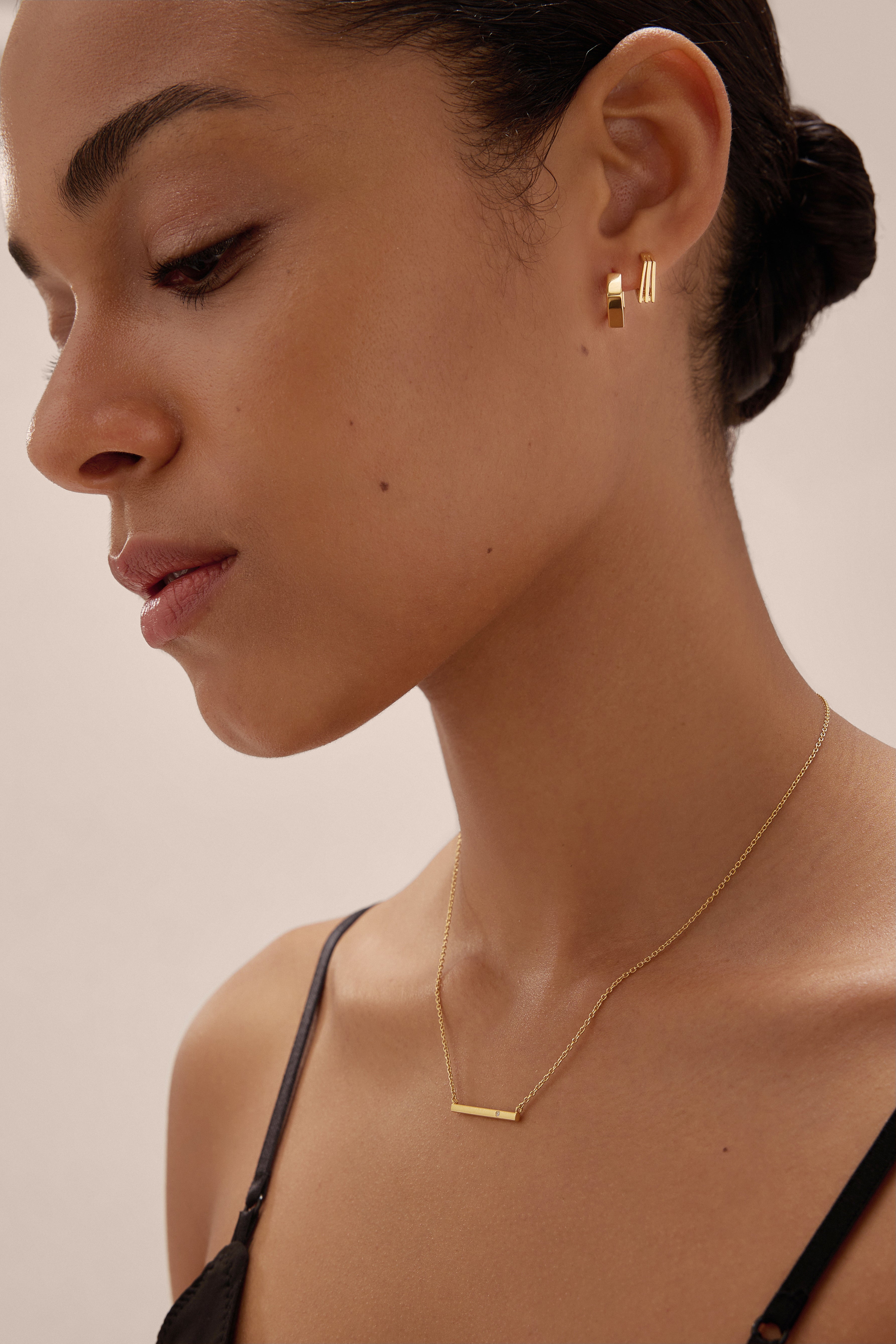 A woman with her dark hair tied back wears the Judith Hexagon Angular Huggies in Gold and a delicate gold bar necklace, dressed in a black strap top, posing against a neutral background.
