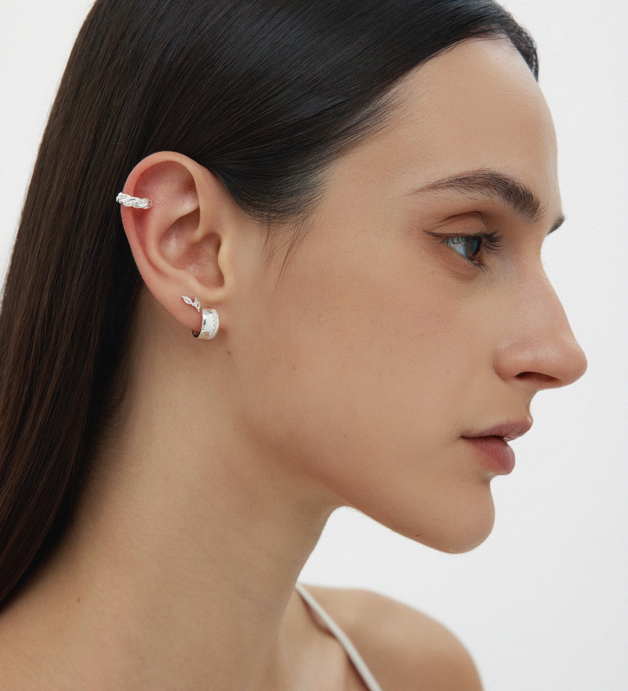 A woman with straight dark hair, in a white thin-strap top, is shown in profile wearing sterling silver ear cuffs and Juni Pavé Chunky Hoop Earrings in Silver on her left ear, against a plain white background.