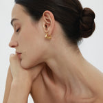 A woman with dark hair in a neat bun poses in profile, eyes closed and chin on her hand. She wears the Juno Ridged Chunky Hoops in Gold and a strapless top against a plain, light background.