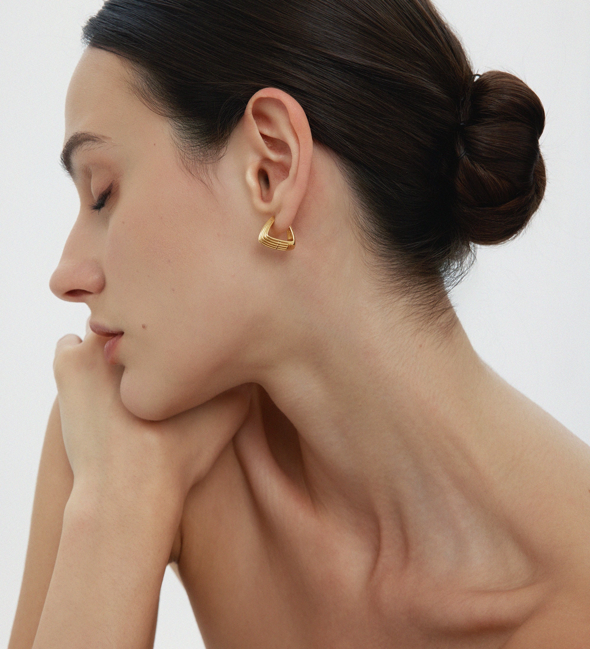 A woman with dark hair in a neat bun poses in profile, eyes closed and chin on her hand. She wears the Juno Ridged Chunky Hoops in Gold and a strapless top against a plain, light background.