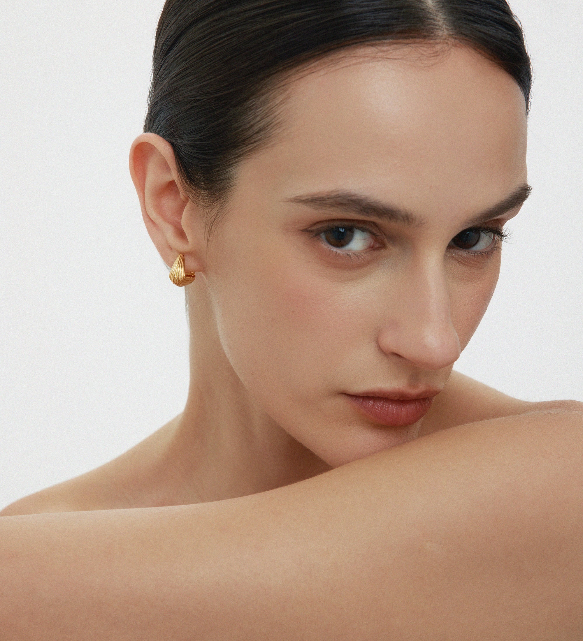 A woman with slicked-back dark hair and bare shoulders gazes at the camera, wearing Juno Ridged Chunky Hoops in Gold and resting her arm across her chest against a plain, light background.