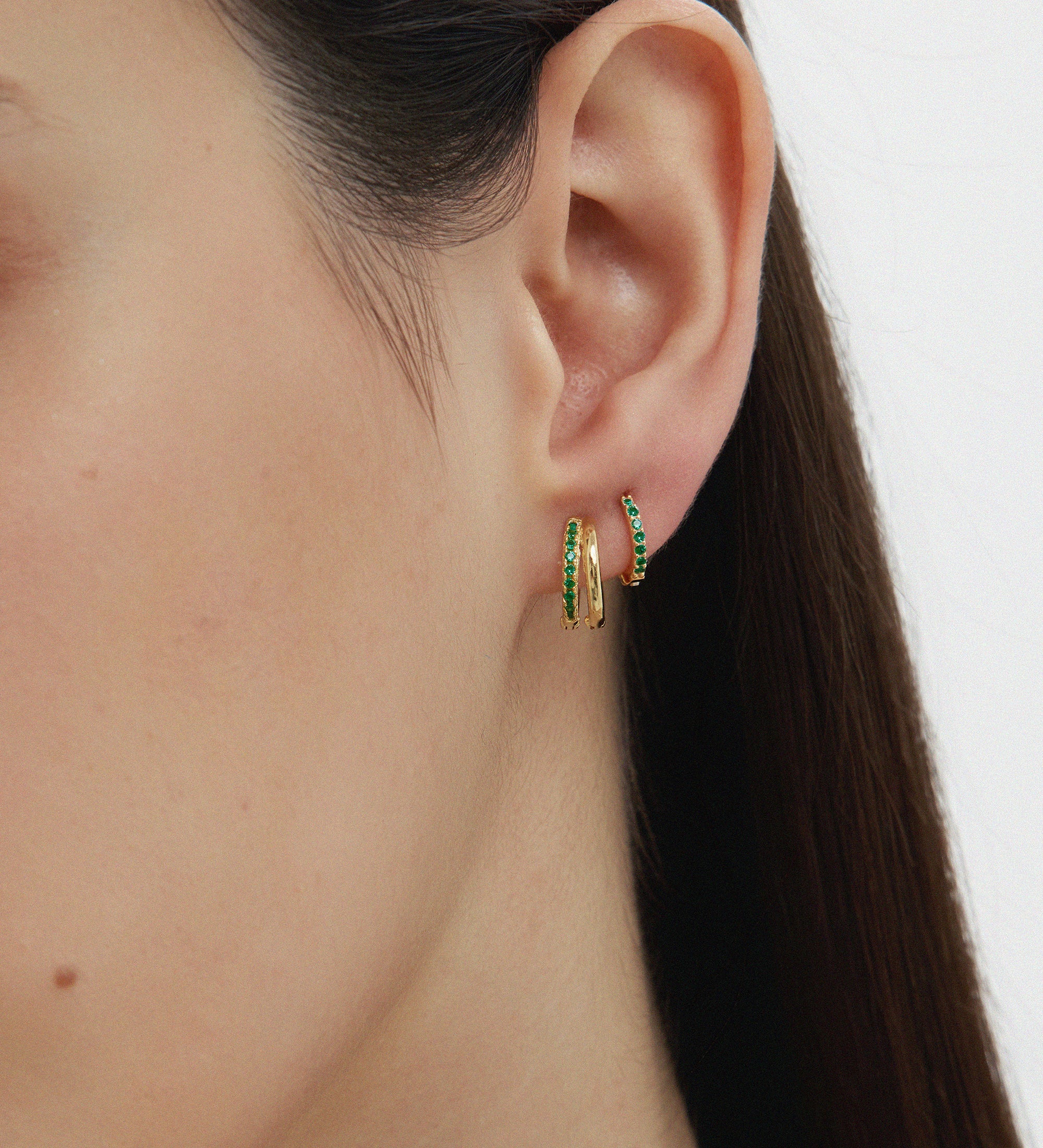Close-up of a womans ear with two gold hoops—one plain, one Kiki Crystal Huggie Earring in Gold/Green. She has straight, dark hair and smooth skin, set against a light background.