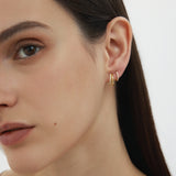 A close-up of a woman with straight dark hair wearing two Kiki Crystal Huggie Earrings in Gold, perfect for ear stacking, in her left ear against a plain light background. Only the left side of her face is visible.