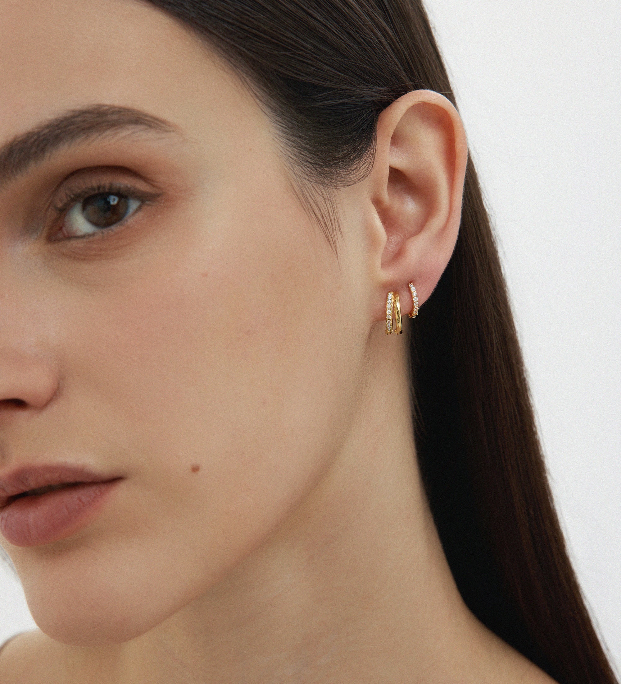 A close-up of a woman with straight dark hair wearing two Kiki Crystal Huggie Earrings in Gold, perfect for ear stacking, in her left ear against a plain light background. Only the left side of her face is visible.