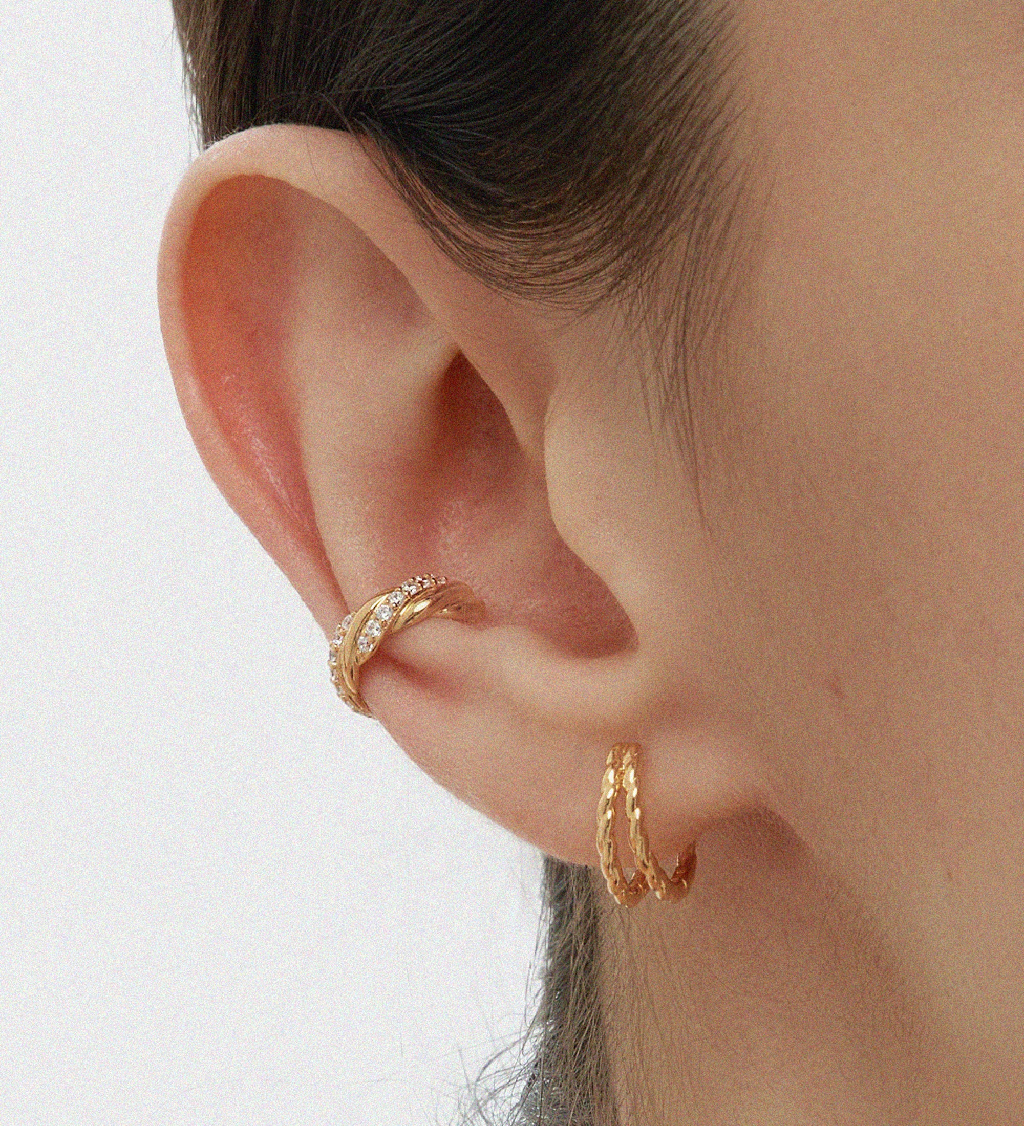 A close-up of a persons ear wearing Mae Twist Double Huggie Earrings in Gold—featuring a textured rope hoop on the lobe and a gold huggie on the cartilage. The background is plain, and hair is tucked behind the ear.