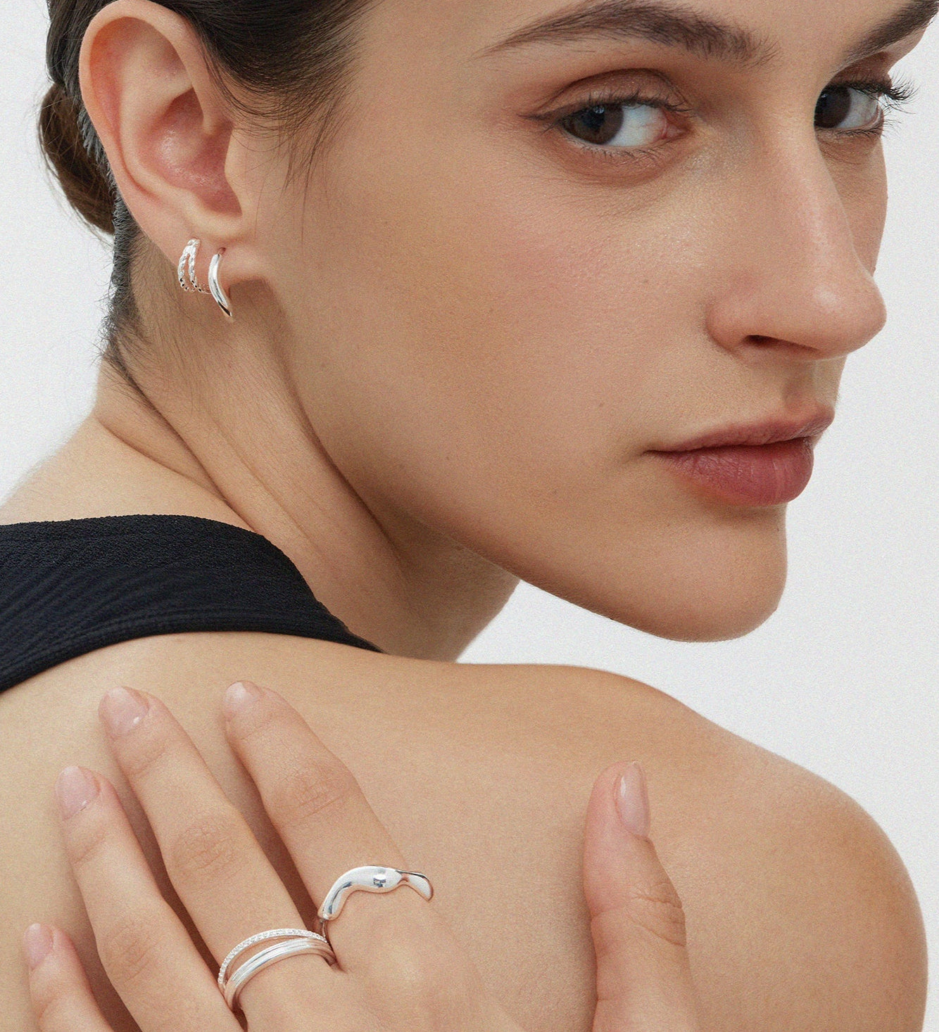 A woman with slicked-back hair wears Mae Twist Double Huggie Earrings in Silver and silver rings, resting her hand on her bare shoulder as she looks over her shoulder at the camera.