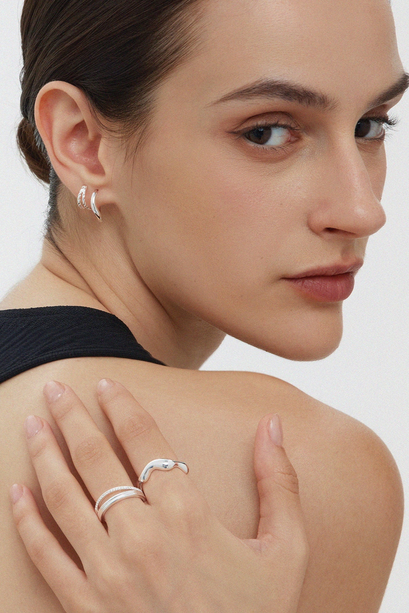 A woman with slicked-back hair wears Mae Twist Double Huggie Earrings in Silver and silver rings, resting her hand on her bare shoulder as she looks over her shoulder at the camera.