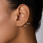 A close-up shows a person’s ear with two 14K Solid Yellow Gold 20G Mini Clickers (6mm and 8mm) in the lobe and a small bow-shaped stud in the upper cartilage, against a neutral background.