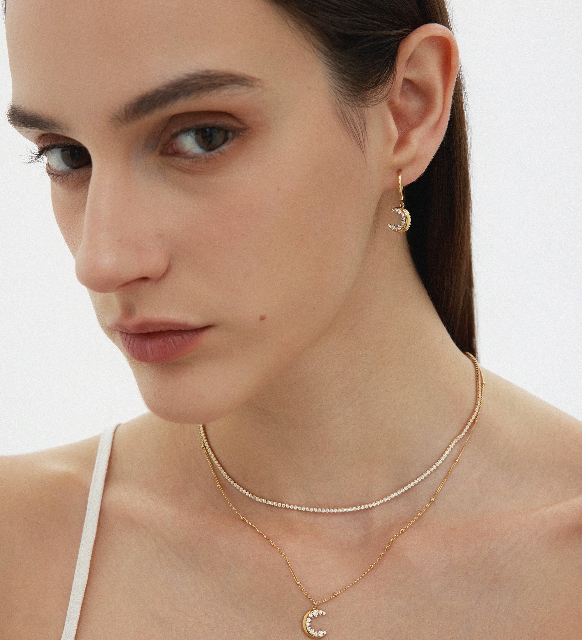 A woman with straight brown hair wears gold jewelry, including the Crescent Moon Crystal Charm Beaded Necklace in Gold. She is dressed in a white spaghetti strap top against a plain background.