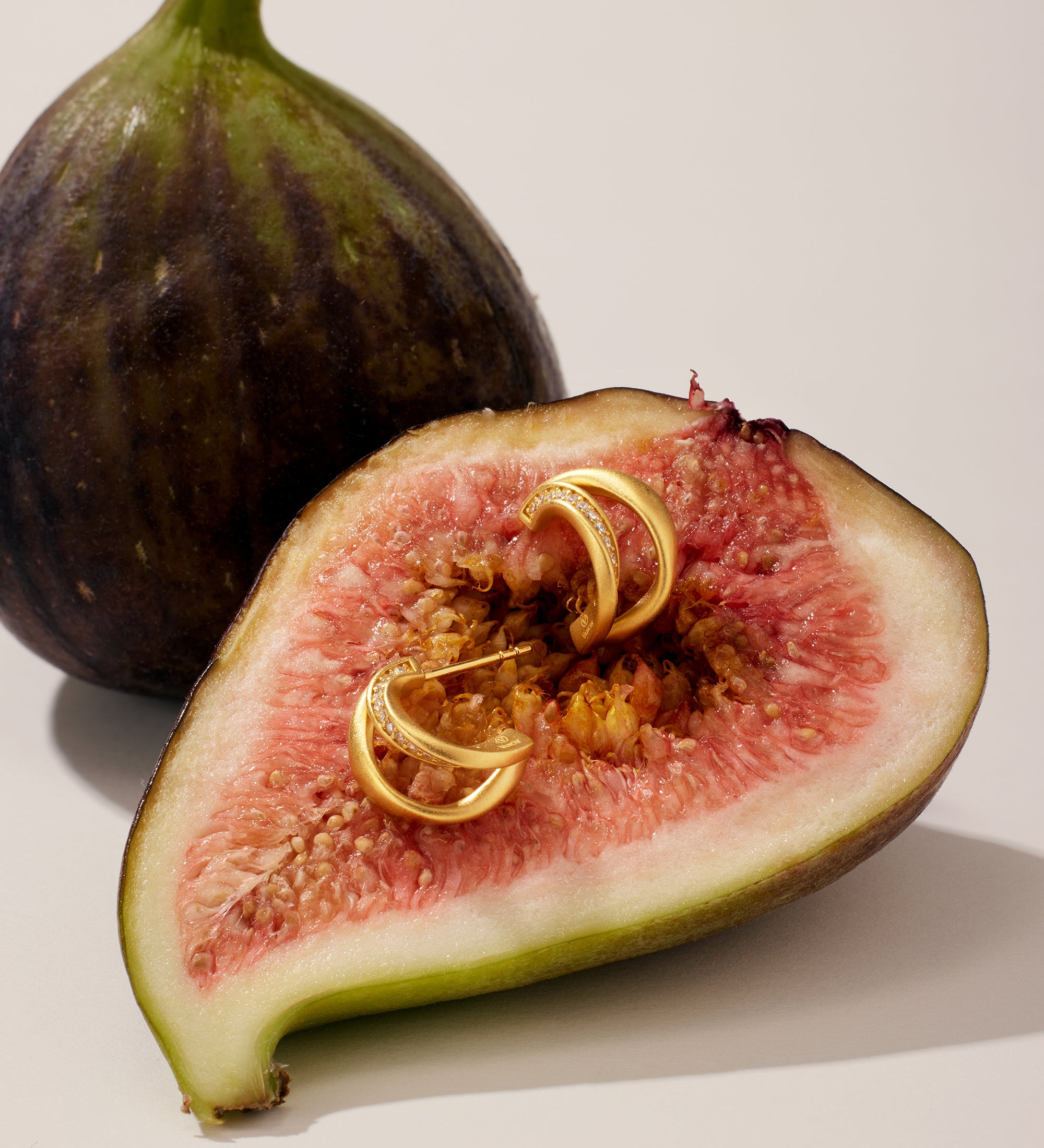 A halved fig with pink flesh holds two Nadine Pavé Twisted Stud Earrings in Gold, while a whole fig stands in the background, both illuminated by soft, natural light.