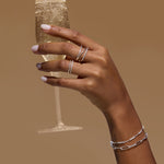 A hand with light pink nail polish wears the Nola Pavé Triple Band Ring in Silver and delicate bracelets while holding a glass of sparkling champagne against a plain brown background.