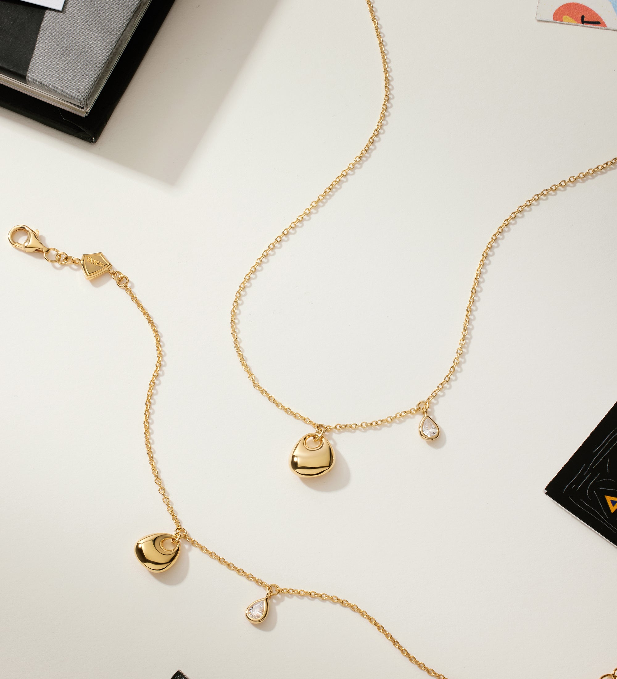 The Nora Double Pendant Bracelet in Gold, featuring sculptural heart and teardrop charms, is displayed on a light surface with scattered books and artistically designed cards.