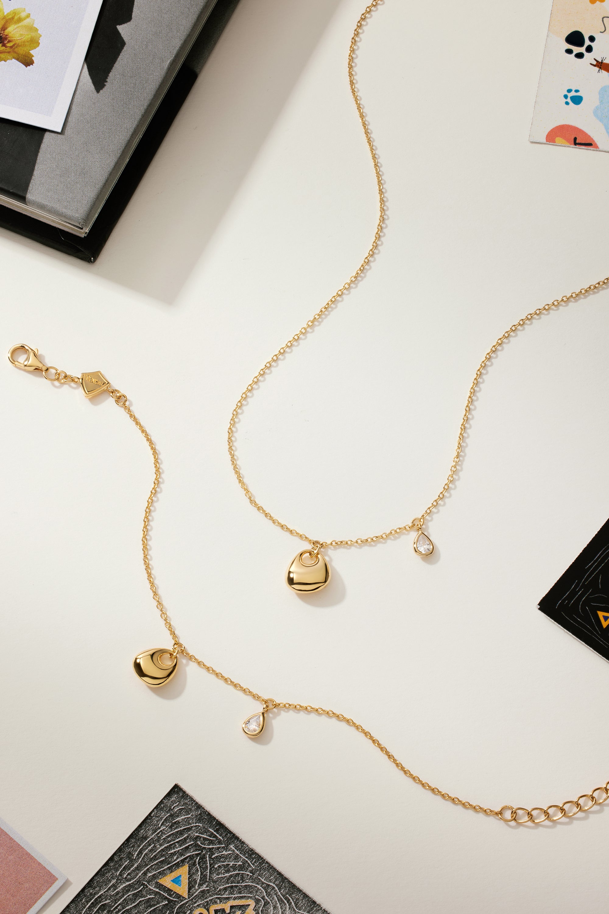 The Nora Double Pendant Bracelet in Gold, featuring sculptural heart and teardrop charms, is displayed on a light surface with scattered books and artistically designed cards.