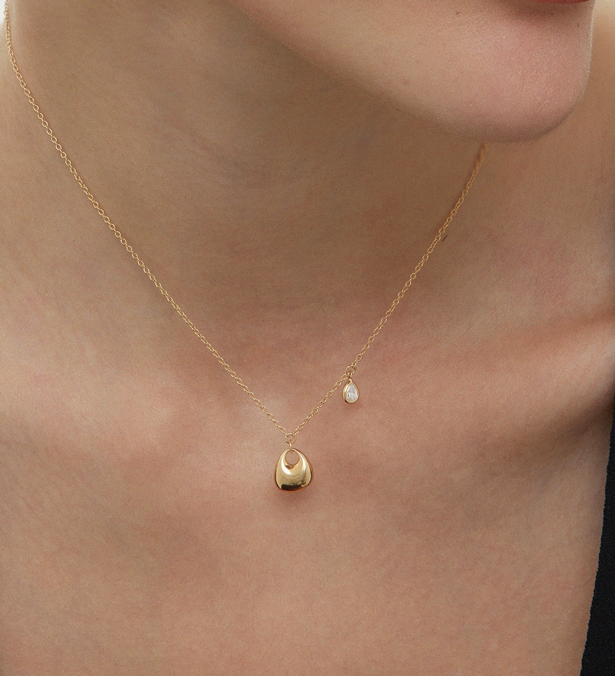 A close-up of a woman’s neck wearing the Nora Double Pendant Necklace in Gold, featuring a small round sculptural pendant and a tiny dangling cubic zirconia, set against light skin and a black top.