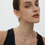 A woman with slicked-back hair wears a black sleeveless top, pearl earrings, and two silver necklaces—one is the Nora Double Pendant Necklace in Silver. She looks confidently at the camera against a plain white background.