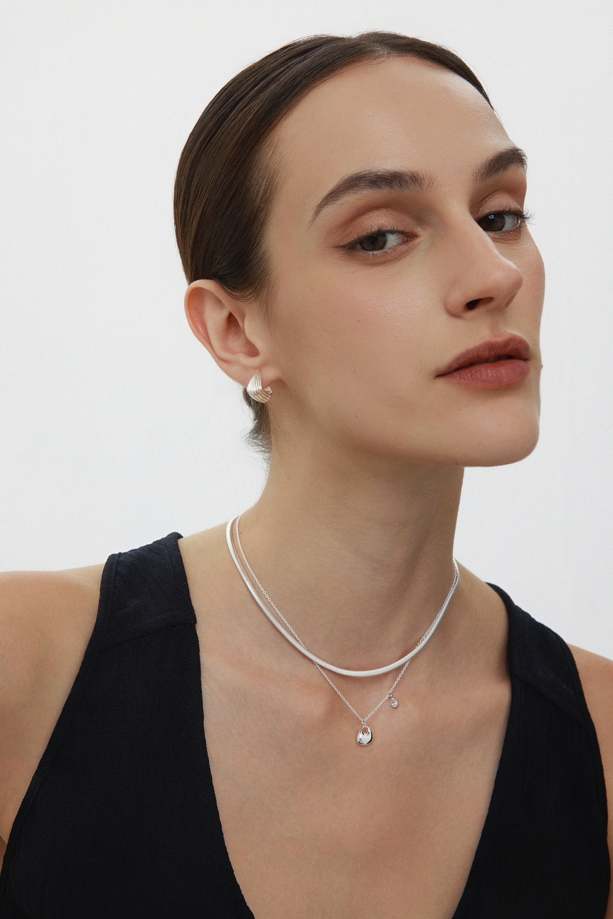 A woman with slicked-back hair wears a black sleeveless top, pearl earrings, and two silver necklaces—one is the Nora Double Pendant Necklace in Silver. She looks confidently at the camera against a plain white background.