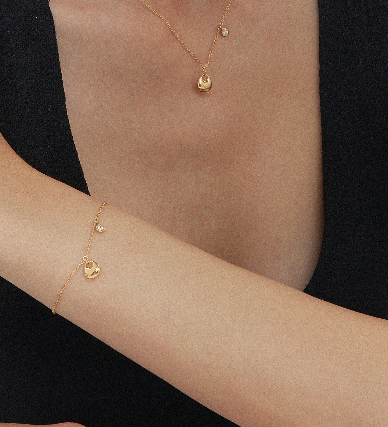 A woman in a sleeveless black top rests her arm on a surface, wearing delicate gold jewelry, including the Nora Double Pendant Necklace in Gold, plus a matching bracelet and ring. Her other hand gently touches her shoulder against a white background.