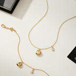 The Nora Double Pendant Necklace in Gold, featuring small sculptural charms such as a teardrop-shaped cubic zirconia, is displayed on a white surface surrounded by scattered colorful papers and cards.