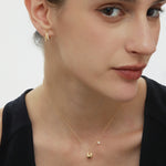 A woman with fair skin and brown hair wears a black sleeveless top, gold earrings, and the Nora Double Pendant Necklace in Gold, posing against a white background.