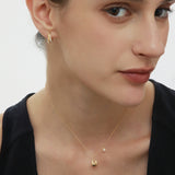 A woman with fair skin and brown hair wears a black sleeveless top, gold earrings, and the Nora Double Pendant Necklace in Gold, posing against a white background.