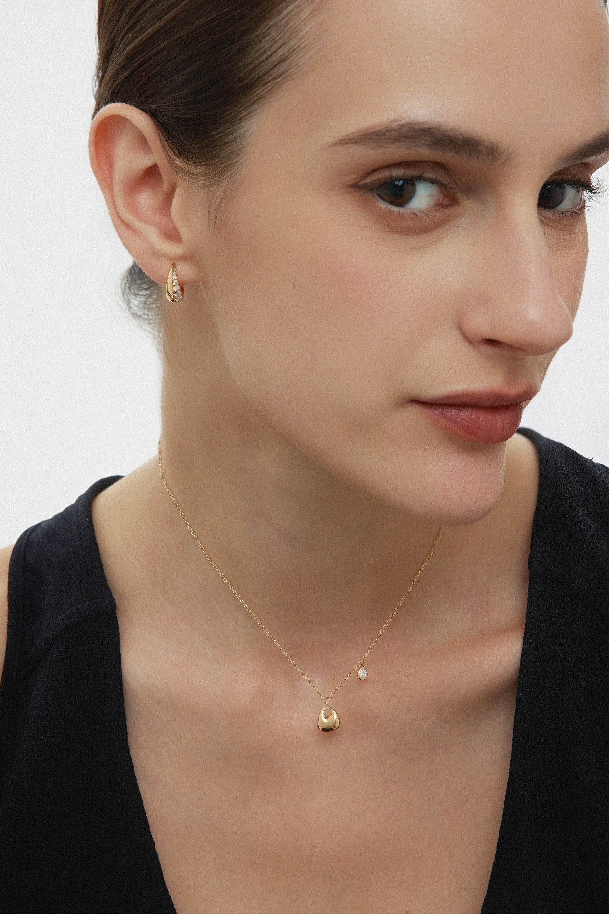 A woman with fair skin and brown hair wears a black sleeveless top, gold earrings, and the Nora Double Pendant Necklace in Gold, posing against a white background.