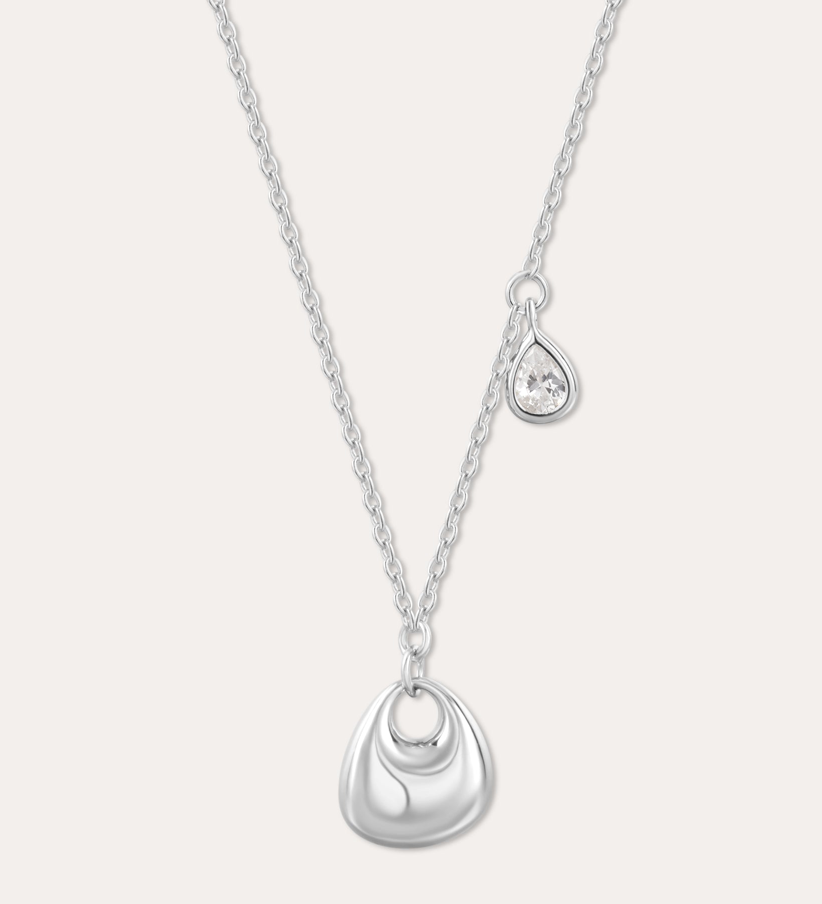 The Nora Double Pendant Necklace in Silver features a fine chain with a shiny sculptural pendant and a separate tiny teardrop charm set with a clear cubic zirconia, displayed against a plain background.