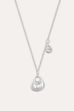 The Nora Double Pendant Necklace in Silver features a fine chain with a shiny sculptural pendant and a separate tiny teardrop charm set with a clear cubic zirconia, displayed against a plain background.