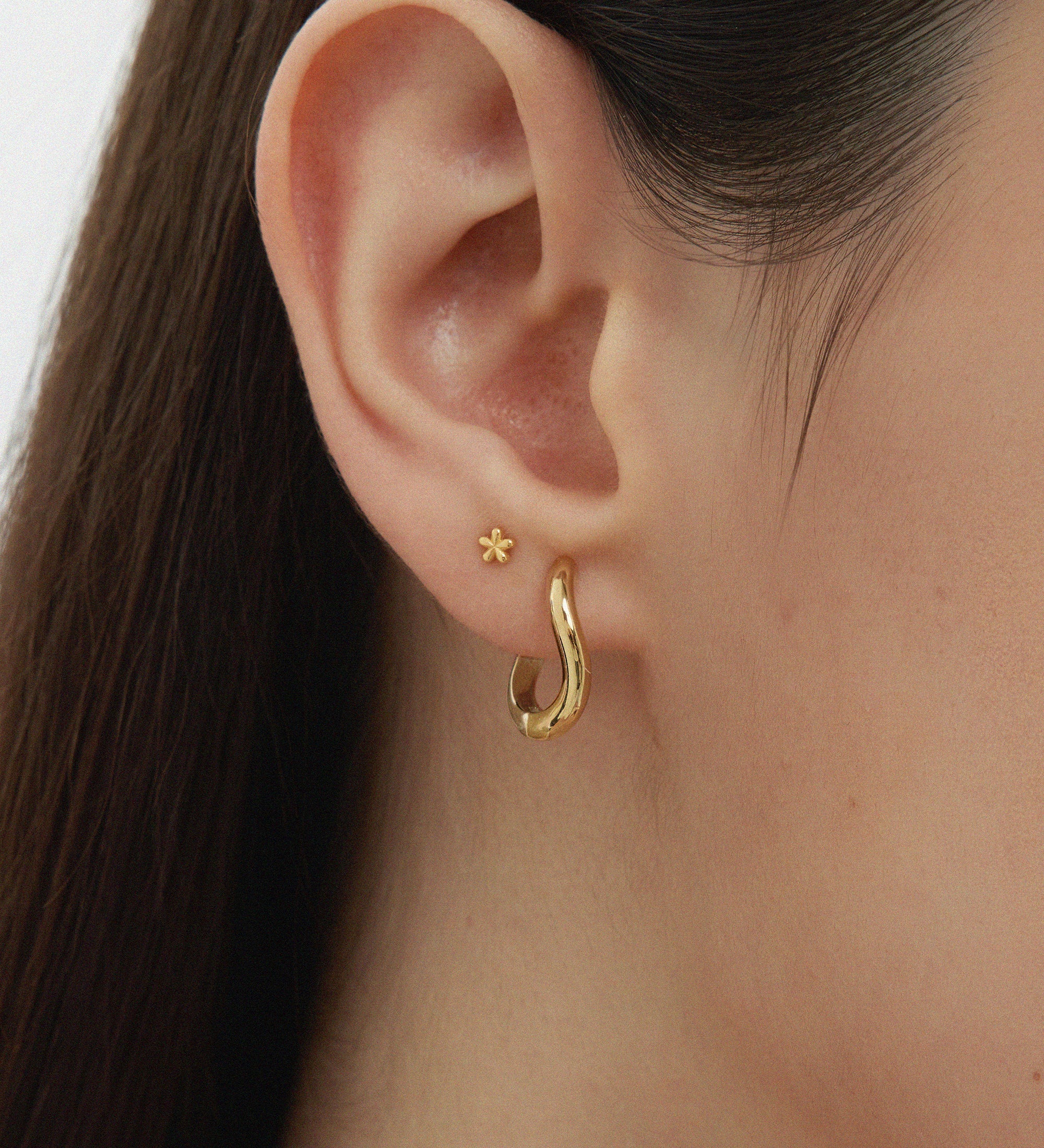 Close-up of a womans ear adorned with two earrings: a small flower stud and the Nori Twisted Hoop Earrings in Gold. The neutral background highlights the elegant shine of these 18k gold plated hoops.