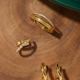 A set of Nori Twisted Hoop Earrings in gold, each featuring elegant twists, is displayed on a wooden surface near a green circular object in the corner.