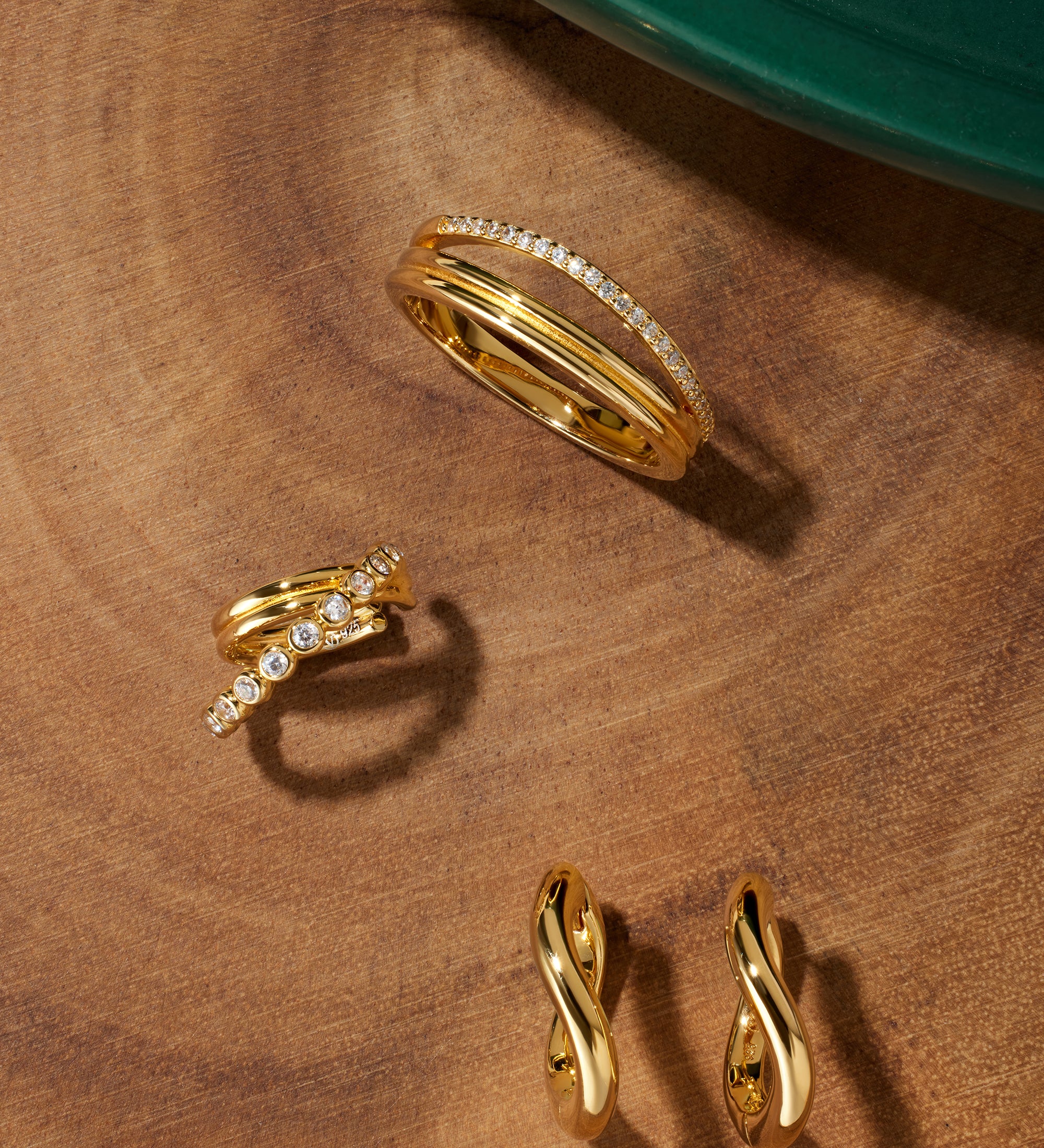 A set of Nori Twisted Hoop Earrings in gold, each featuring elegant twists, is displayed on a wooden surface near a green circular object in the corner.