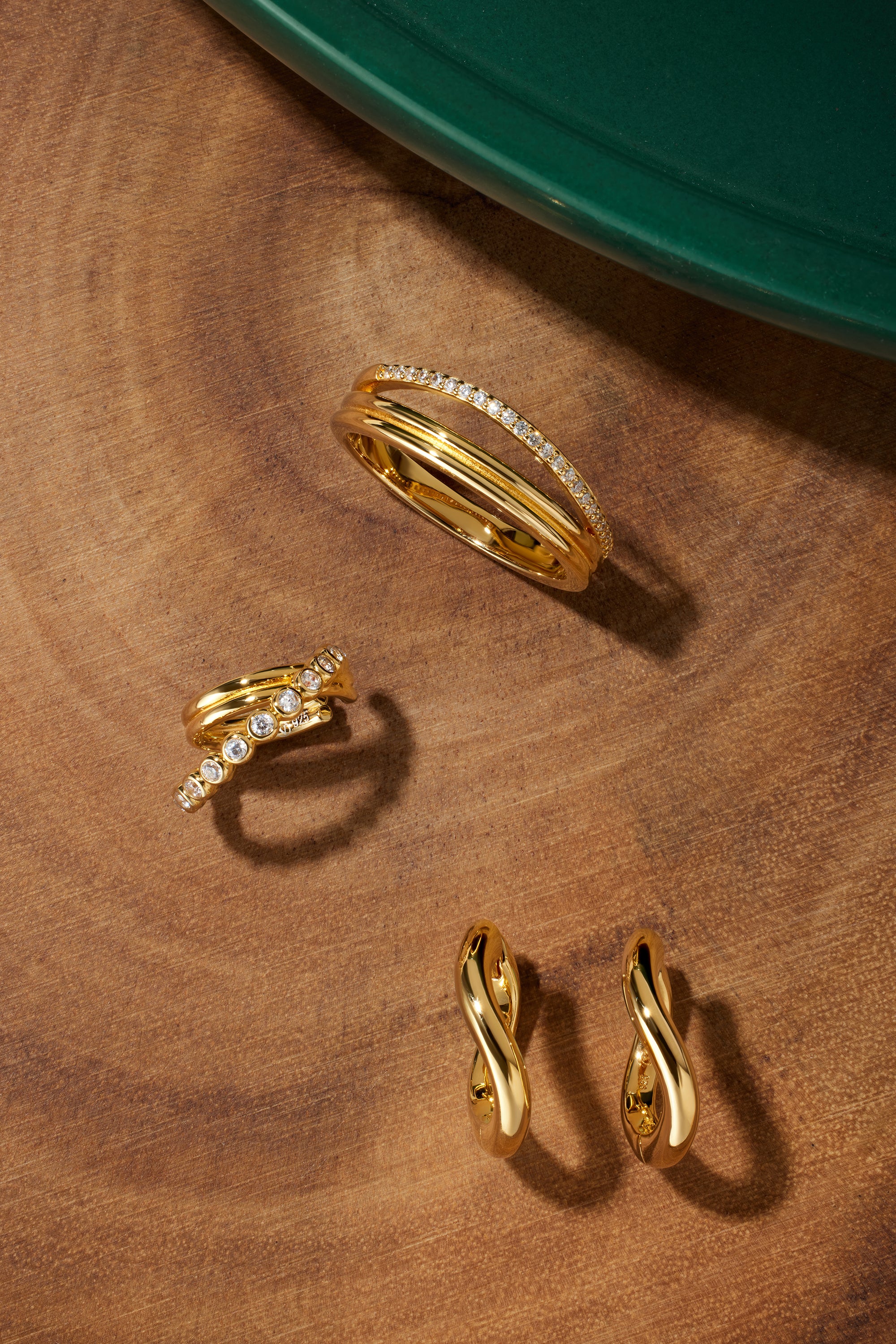 A set of Nori Twisted Hoop Earrings in gold, each featuring elegant twists, is displayed on a wooden surface near a green circular object in the corner.