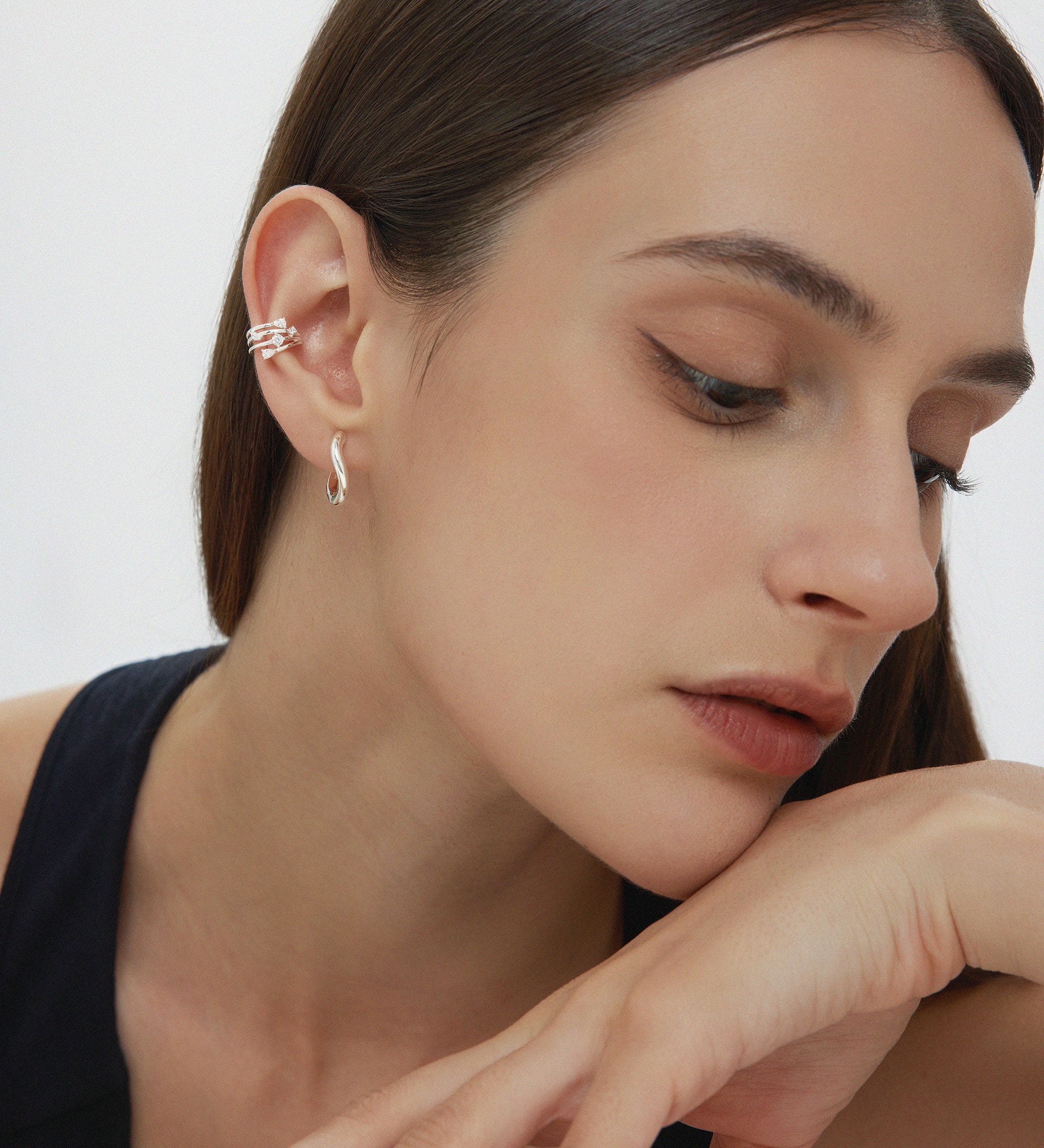 A woman with straight brown hair rests her chin on her hand, wearing Nori Twisted Hoop Earrings in Silver and stacked ear cuffs. She has natural makeup and a sleeveless black top, posed against a plain background.
