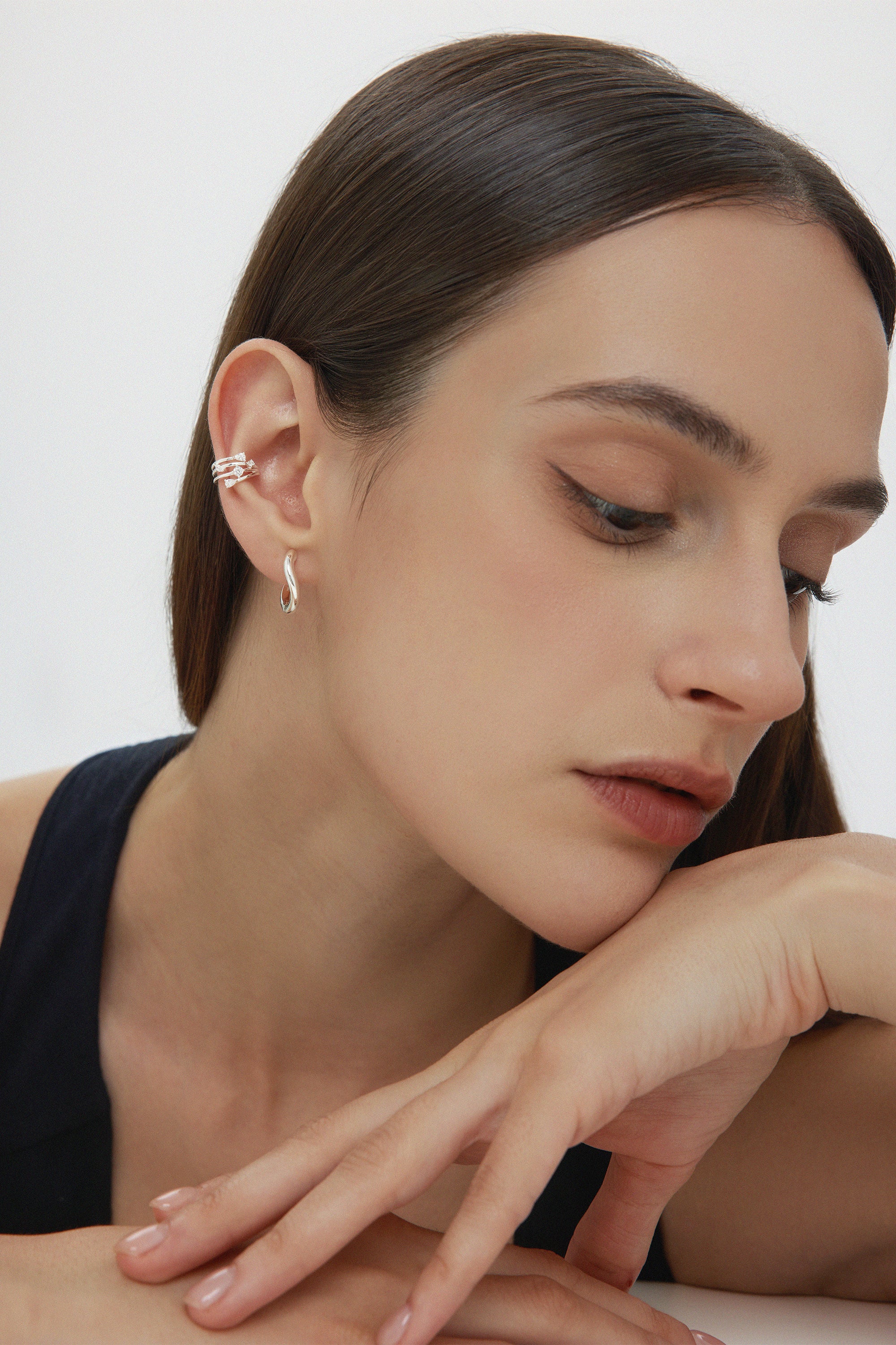 A woman with straight brown hair rests her chin on her hand, wearing Nori Twisted Hoop Earrings in Silver and stacked ear cuffs. She has natural makeup and a sleeveless black top, posed against a plain background.