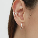 Close-up of a person’s ear with straight brown hair, wearing sterling silver hoop earrings, including the Nori Twisted Hoop Earrings in Silver in the earlobe and a crystal-studded silver ear cuff on the upper ear.