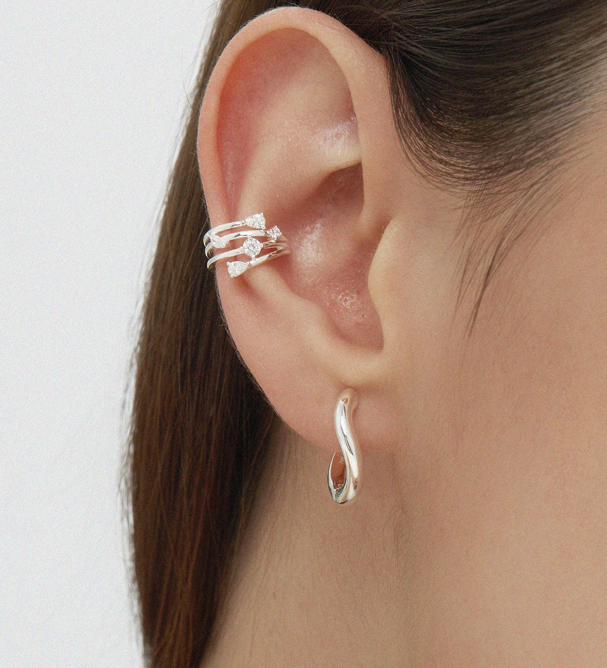 Close-up of a person’s ear with straight brown hair, wearing sterling silver hoop earrings, including the Nori Twisted Hoop Earrings in Silver in the earlobe and a crystal-studded silver ear cuff on the upper ear.