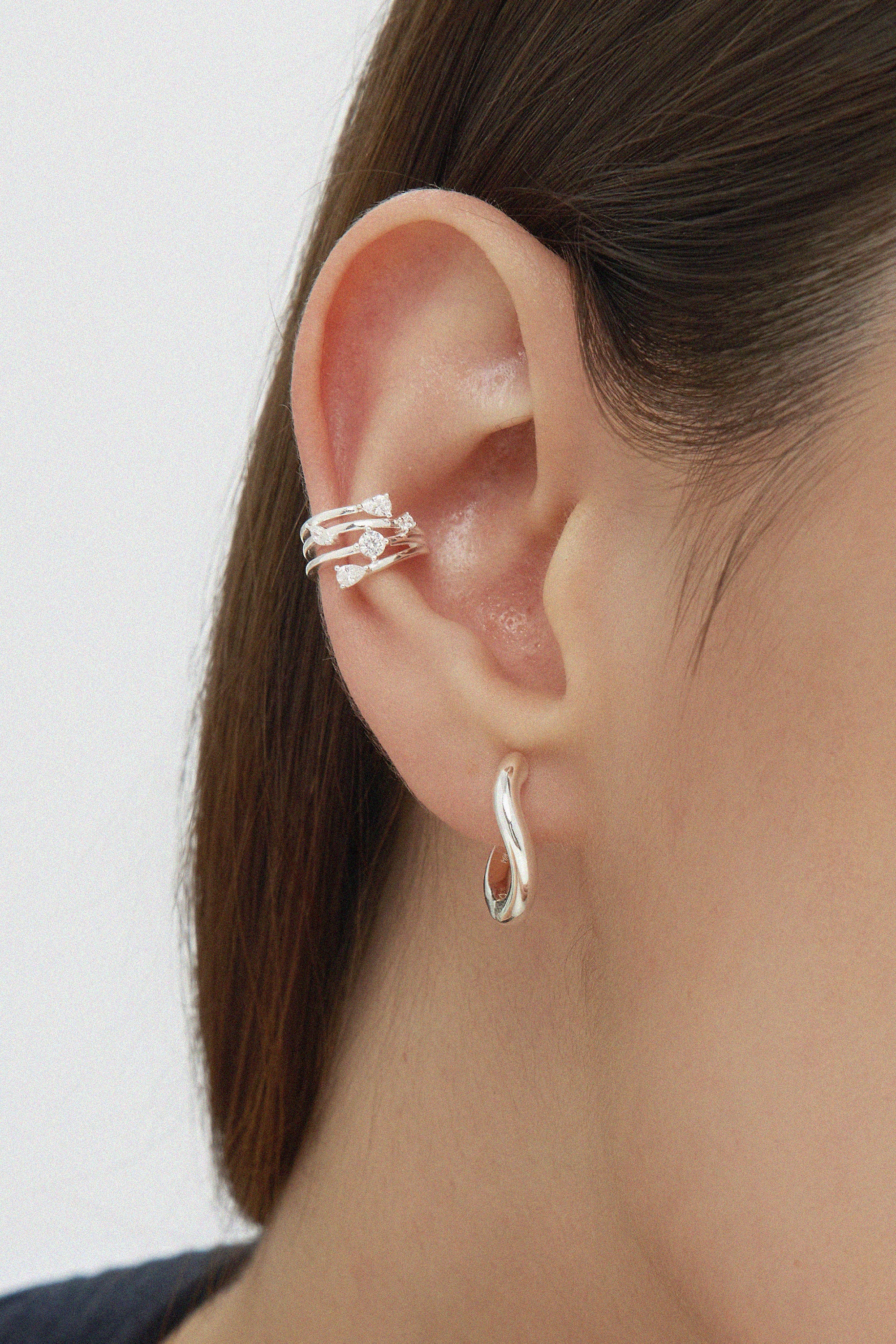 Close-up of a person’s ear with straight brown hair, wearing sterling silver hoop earrings, including the Nori Twisted Hoop Earrings in Silver in the earlobe and a crystal-studded silver ear cuff on the upper ear.