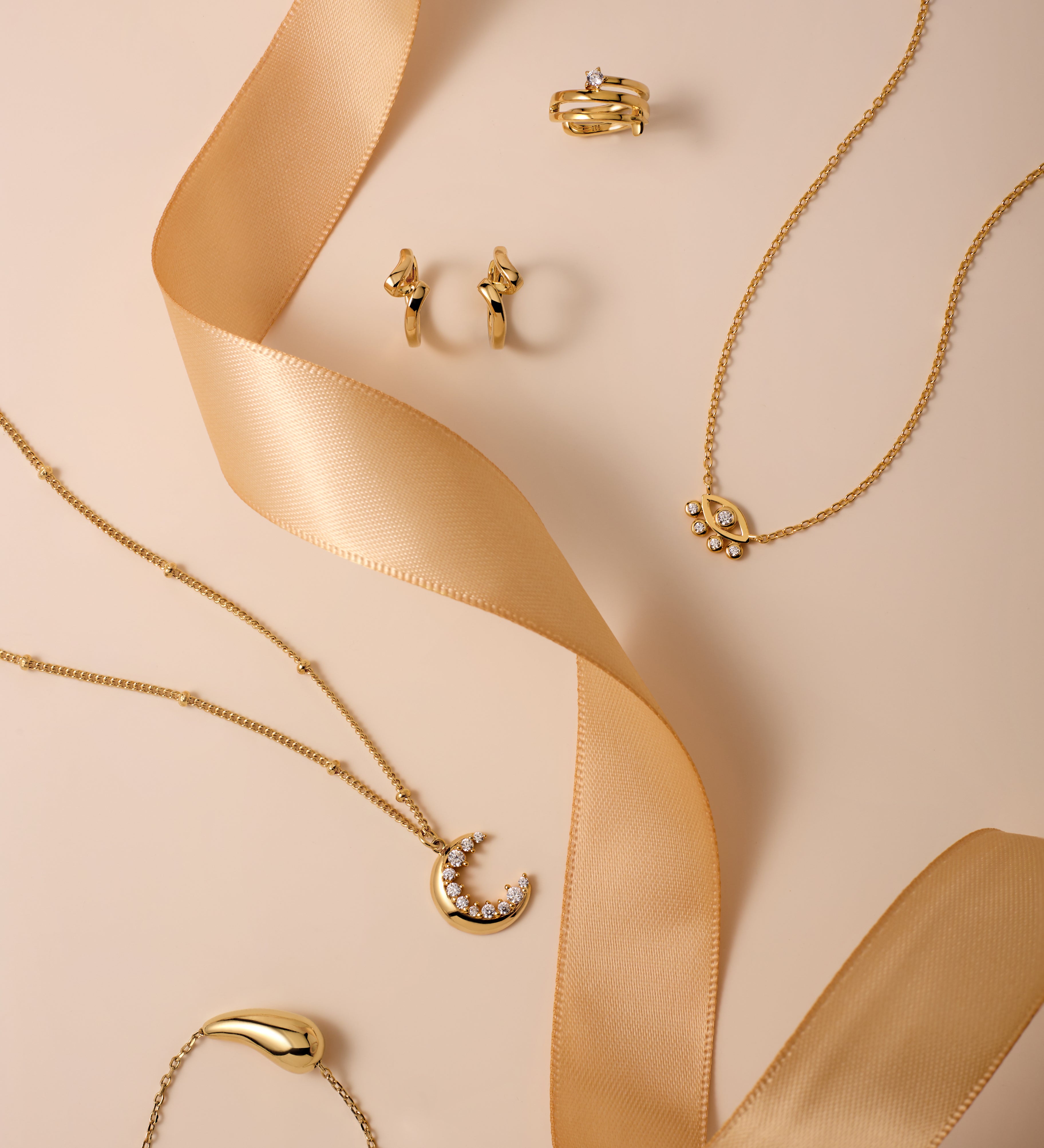 Gold Ribbon Twist Huggies are displayed on a beige surface with a satin ribbon, alongside gold necklaces featuring crescent moon pendants and delicate chains for an elegant, stylish look.