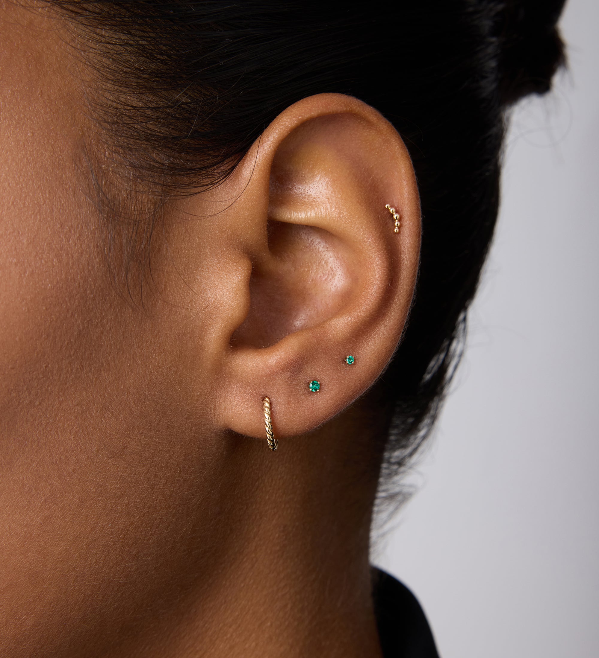 A close-up of an ear with four piercings displays small sustainable gold earrings, turquoise studs, and a 14K Recycled Solid Yellow Gold Rope Huggie Earring. The person has dark hair pulled back and wears a black top.