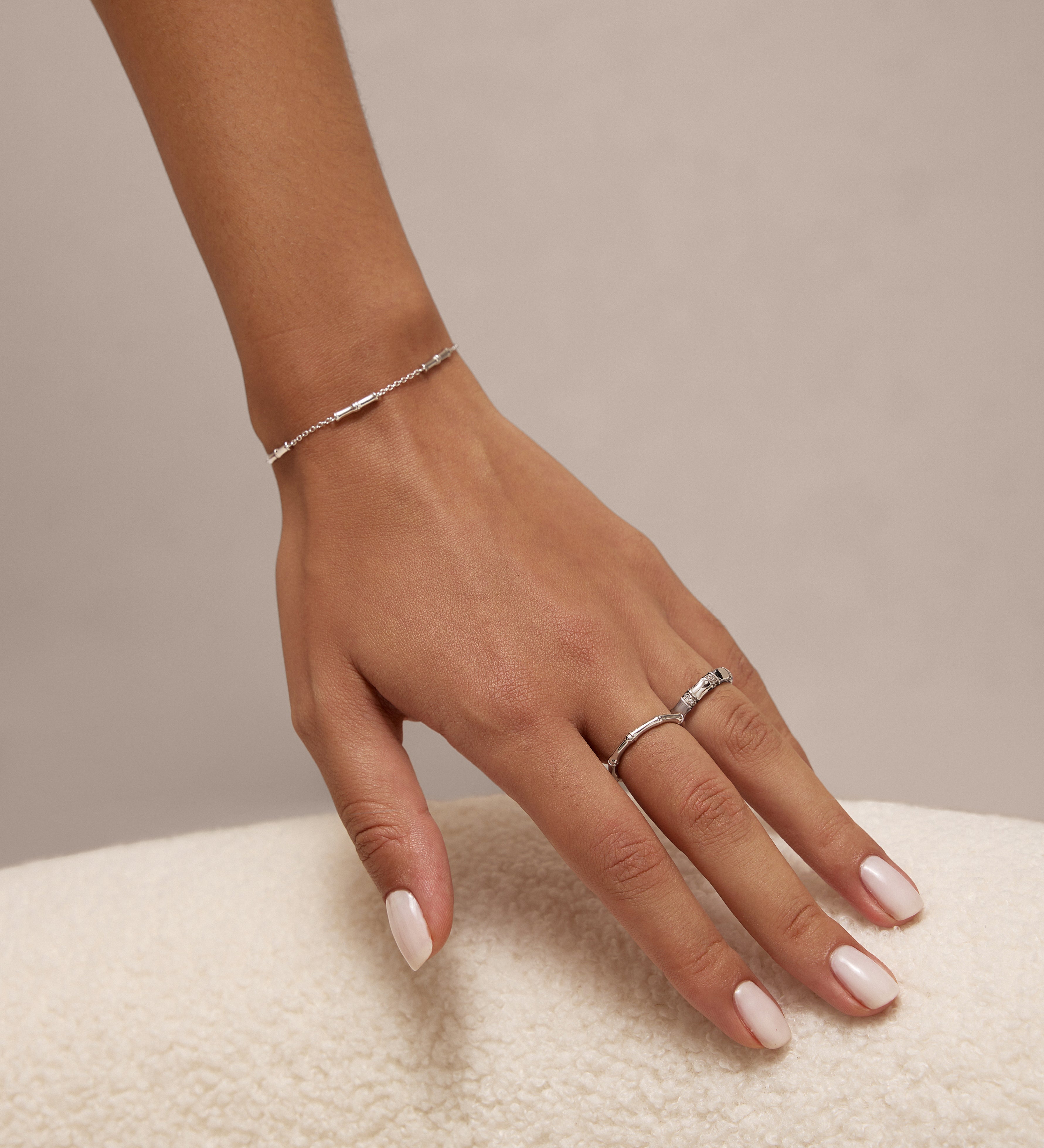 A close-up of a womans light pink manicured hand wearing the Bamboo Ring in Silver—a minimalist piece crafted from recycled sterling silver—plus another simple ring and a delicate silver bracelet, resting on a textured white surface.