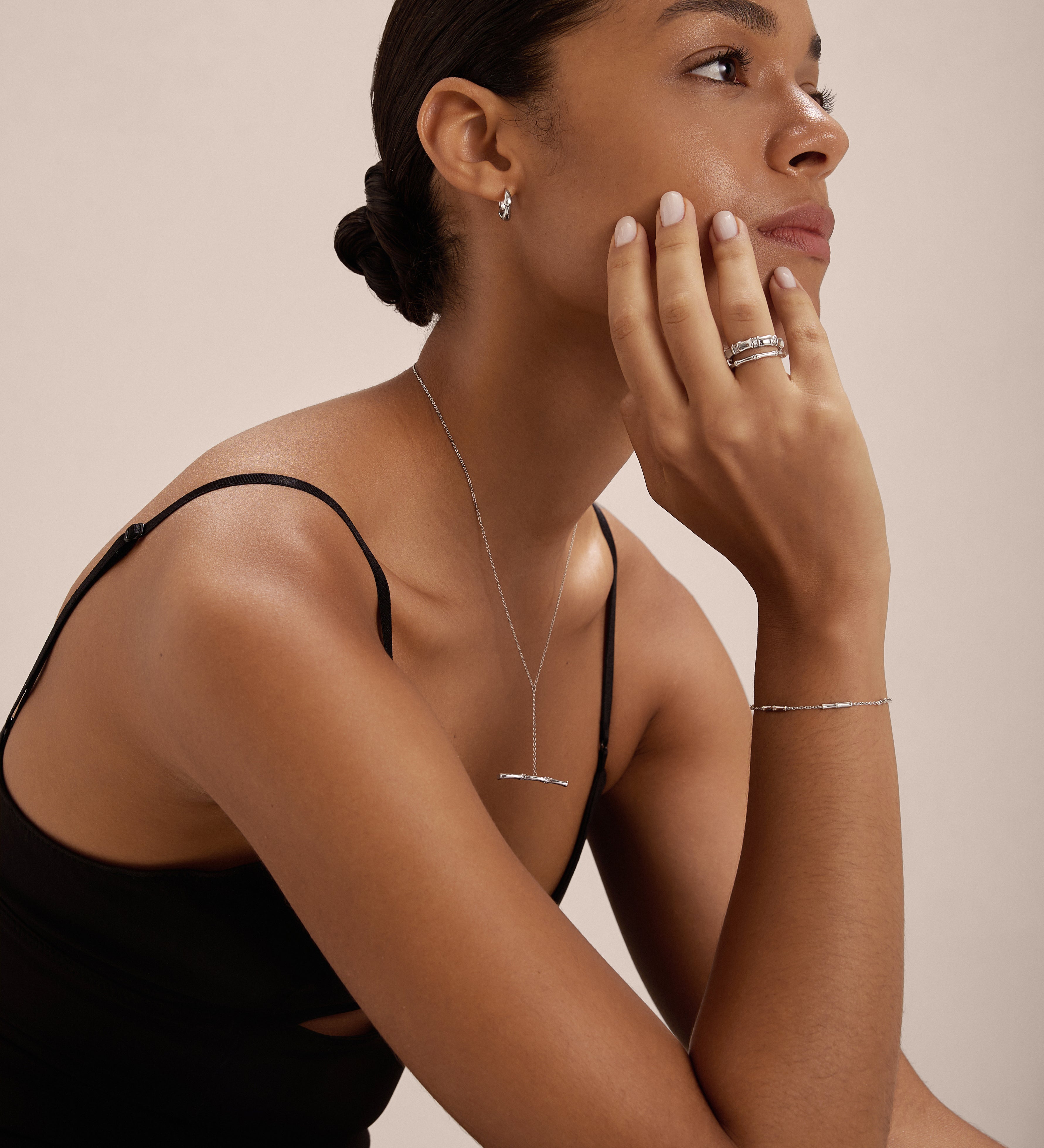 A woman with dark hair in a low bun wears the Colette Droop Link Huggie Earrings in Silver, a black sleeveless top, and other silver jewelry as she rests her chin on her hand and gazes to the side against a plain background.