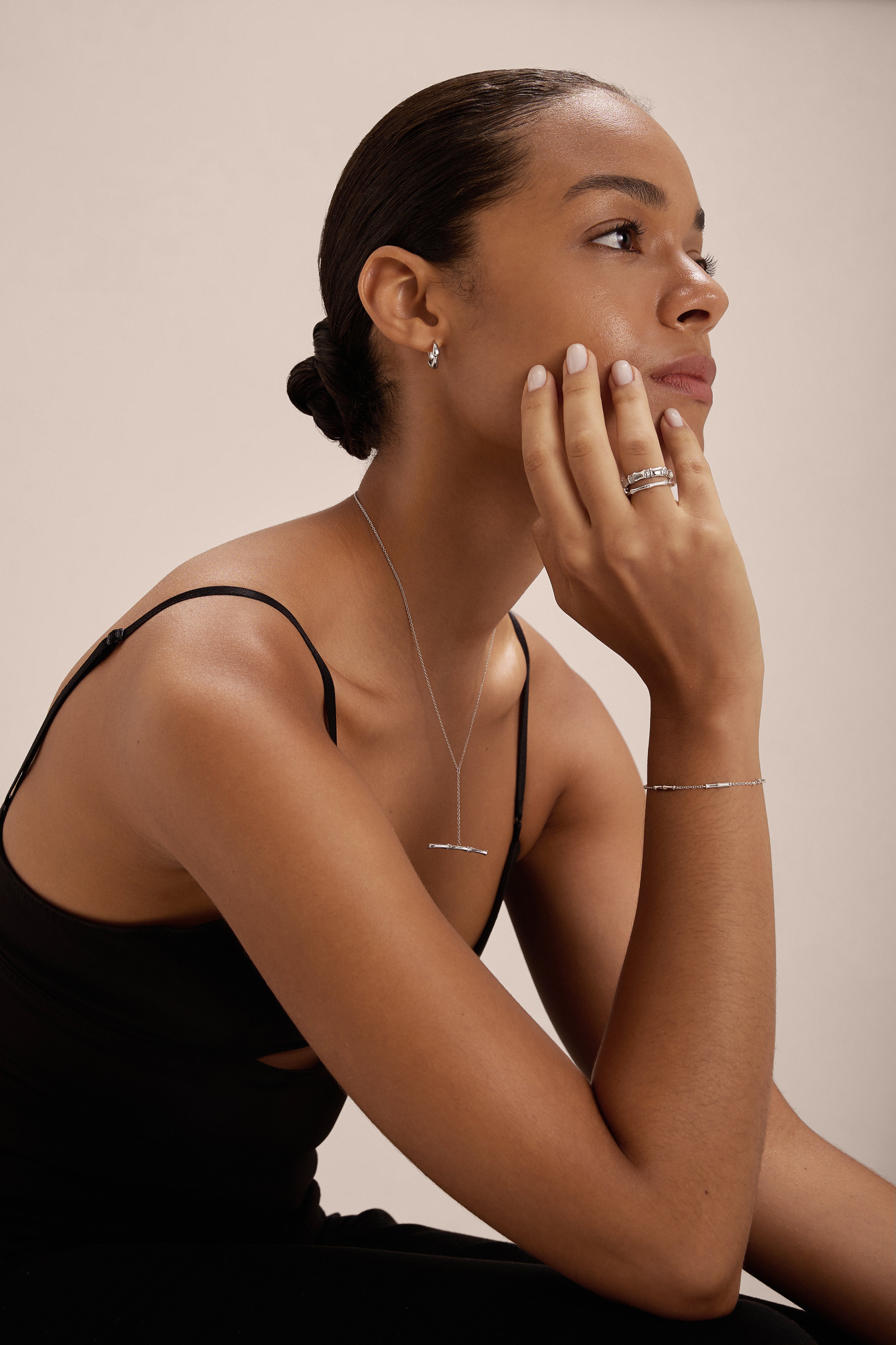A woman with dark hair in a low bun wears the Colette Droop Link Huggie Earrings in Silver, a black sleeveless top, and other silver jewelry as she rests her chin on her hand and gazes to the side against a plain background.